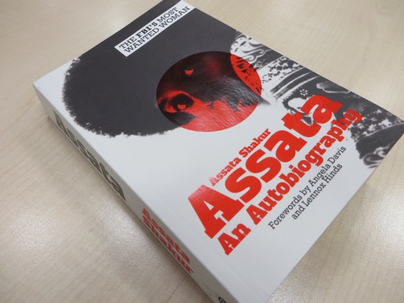 the picture shows a book on a table. The book cover has a young black woman's face in profile, with a red target on her face. The title is Assata: An Autobiography