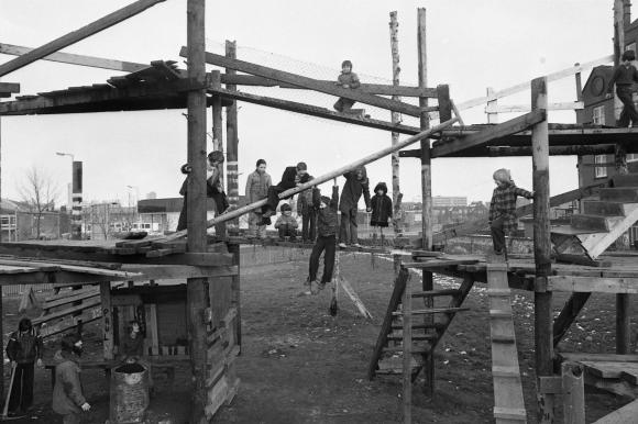 A black and white photograph of several children balancing on plank connecting two parts of an adventure playground